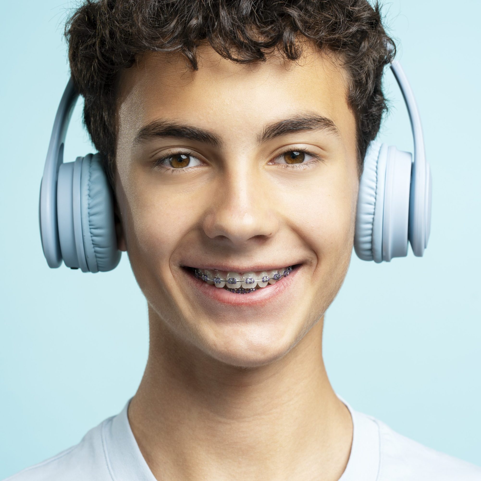 Closeup portrait of smiling handsome teenage boy in dental braces wearing headphones listening to music isolated on blue background. Advertisement concept, technology